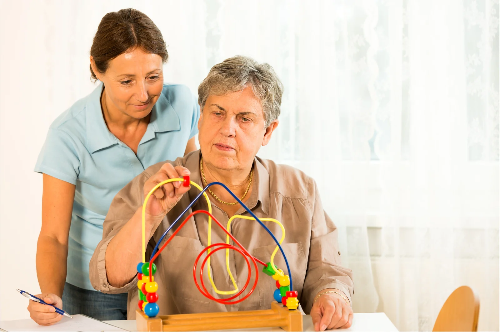 Caregiver observing senior woman doing cognitive therapy with a colorful bead maze