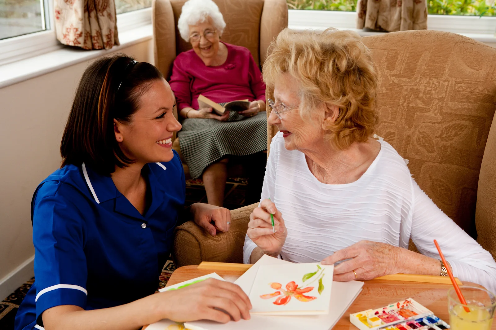 Caregiver assisting senior woman with watercolor painting in a comfortable living room