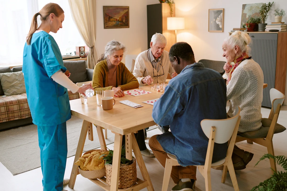Caregiver supervising seniors playing cards at a table in a cozy common room
