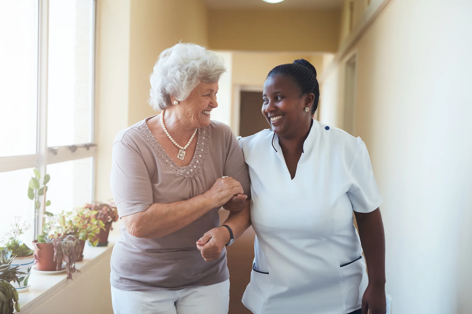 Senior woman and caregiver walking arm in arm down a sunny hallway