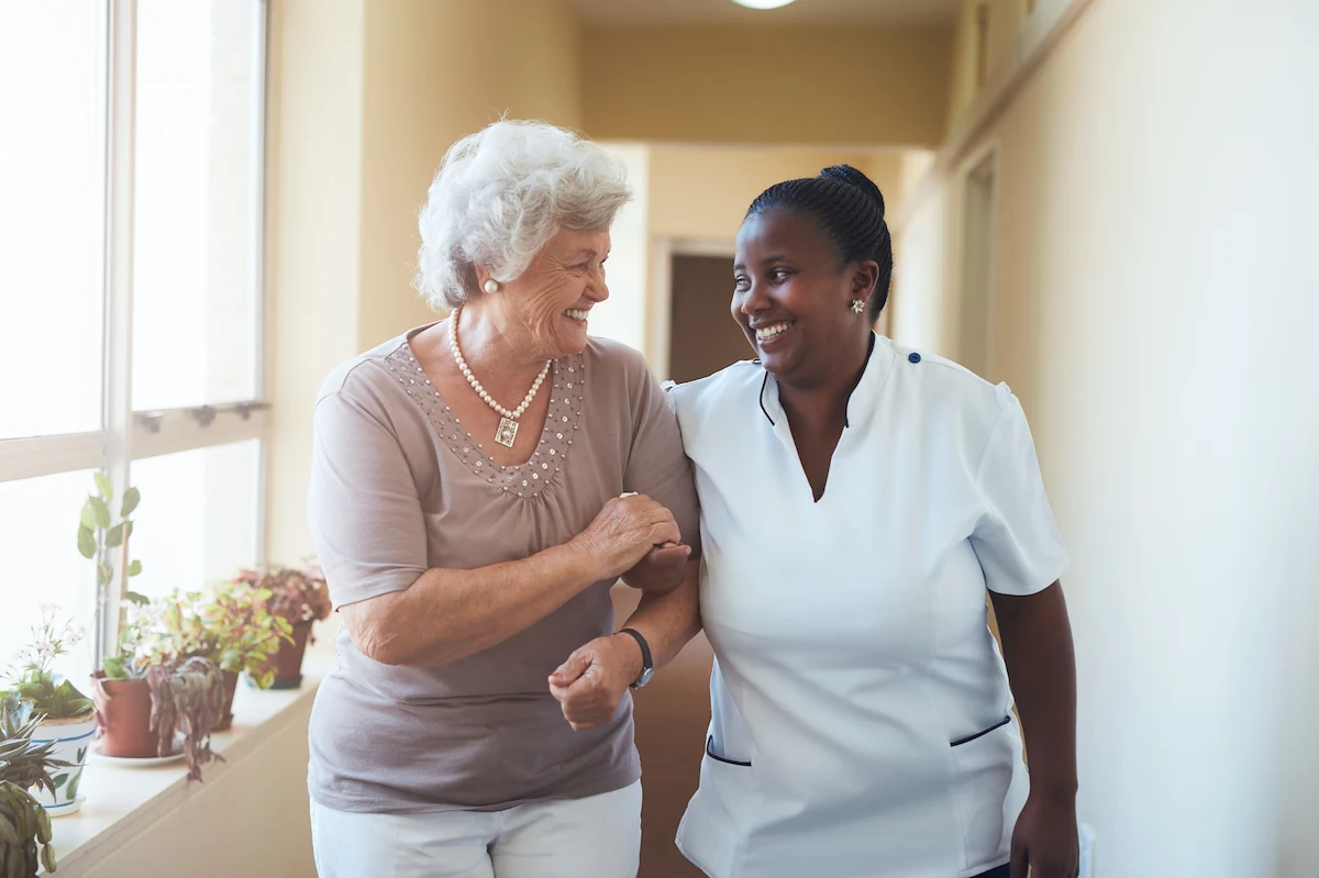 Senior woman and caregiver walking arm in arm down a sunny hallway