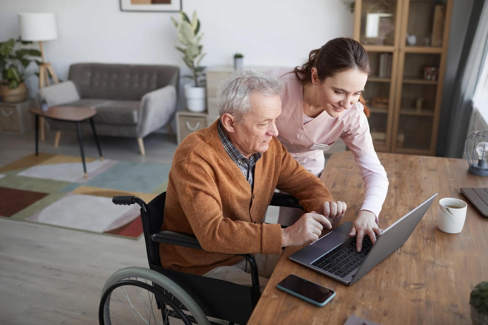 Caregiver helping senior man in wheelchair use a laptop at home