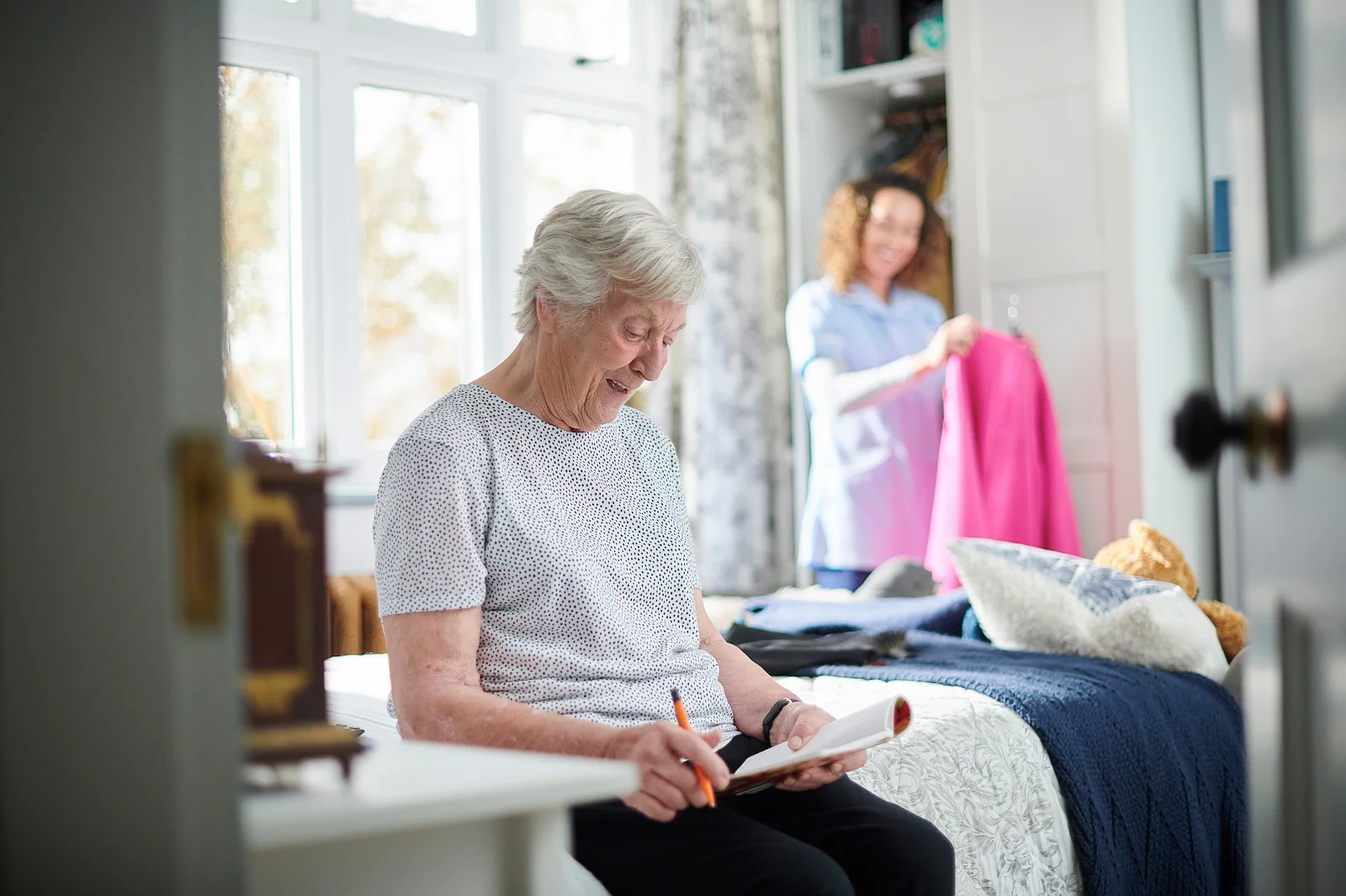 Senior woman reading while caregiver organizes laundry in the bedroom