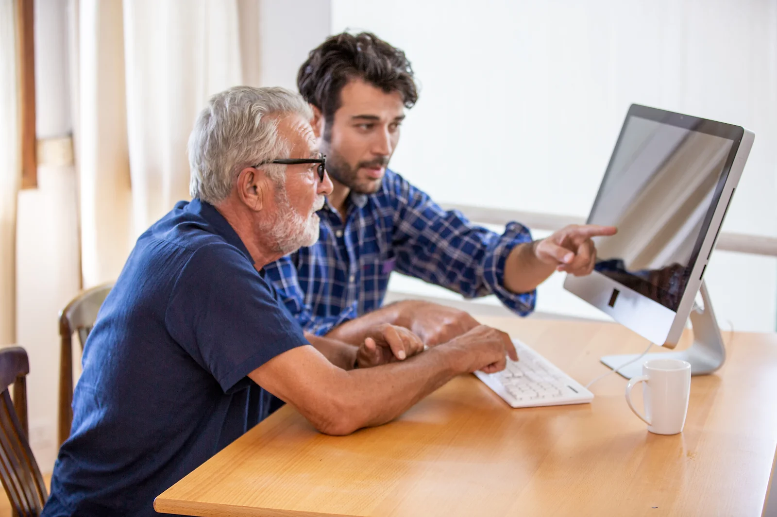 Young man helping senior man navigate a computer at the dining table