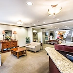 A spacious, warmly lit lobby in Godfrey, Illinois, with striped sofas, a wooden coffee table, and a granite reception desk—perfect for memory care or assisted living. Decorative lamps, plants, mirrors, and photos enhance the welcoming atmosphere.