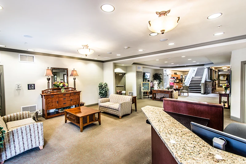 A spacious, warmly lit lobby in Godfrey, Illinois, with striped sofas, a wooden coffee table, and a granite reception desk—perfect for memory care or assisted living. Decorative lamps, plants, mirrors, and photos enhance the welcoming atmosphere.