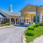 Exterior view of Charter Senior Living of Hermitage in Hermitage, TN, featuring a brick building with yellow accents, drive-through drop-off area, and rocking chairs on the front porch, offering independent living, assisted living, and memory care.