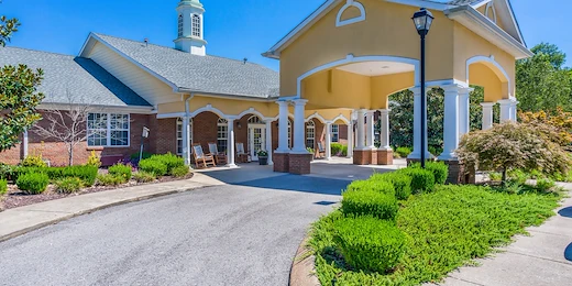 Exterior view of Charter Senior Living of Hermitage in Hermitage, TN, featuring a brick building with yellow accents, drive-through drop-off area, and rocking chairs on the front porch, offering independent living, assisted living, and memory care.
