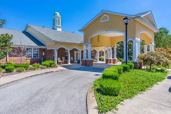 Exterior view of Charter Senior Living of Hermitage in Hermitage, TN, featuring a brick building with yellow accents, drive-through drop-off area, and rocking chairs on the front porch, offering independent living, assisted living, and memory care.