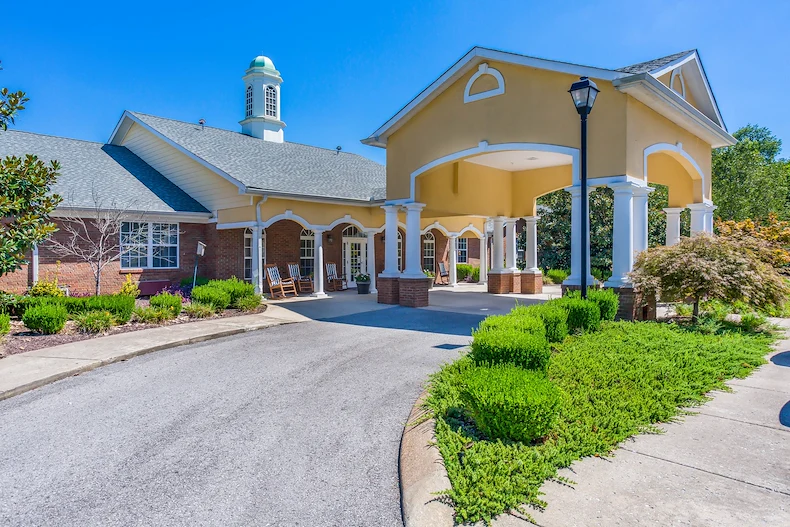 Exterior view of Charter Senior Living of Hermitage in Hermitage, TN, featuring a brick building with yellow accents, drive-through drop-off area, and rocking chairs on the front porch, offering independent living, assisted living, and memory care.