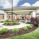 Charter Senior Living of Godfrey is a large brick building with a covered driveway entrance, surrounded by landscaped gardens and green lawns. A person enjoys the outdoors on a bench under the canopy with their dog—perfect for assisted living or memory care.
