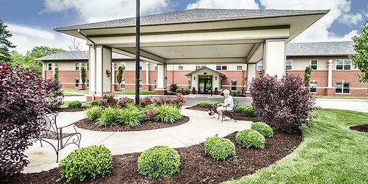 Charter Senior Living of Godfrey is a large brick building with a covered driveway entrance, surrounded by landscaped gardens and green lawns. A person enjoys the outdoors on a bench under the canopy with their dog—perfect for assisted living or memory care.