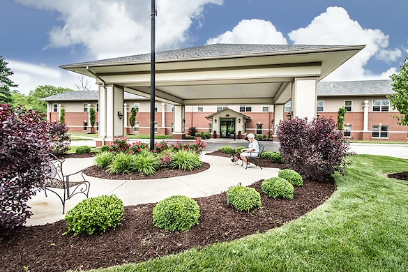 Charter Senior Living of Godfrey is a large brick building with a covered driveway entrance, surrounded by landscaped gardens and green lawns. A person enjoys the outdoors on a bench under the canopy with their dog—perfect for assisted living or memory care.