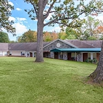 Exterior of Charter Senior Living of Jackson in Jackson, TN, showcasing a brick building with a covered awning extension and a large grassy area with mature trees, providing a welcoming environment for assisted living and memory care residents.