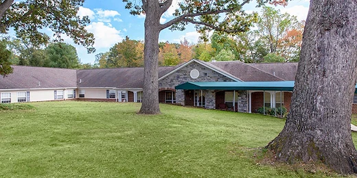 Exterior of Charter Senior Living of Jackson in Jackson, TN, showcasing a brick building with a covered awning extension and a large grassy area with mature trees, providing a welcoming environment for assisted living and memory care residents.