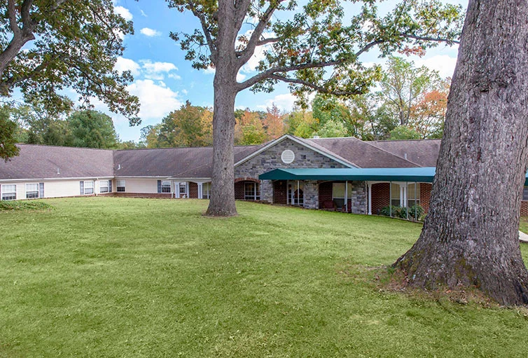 Exterior of Charter Senior Living of Jackson in Jackson, TN, showcasing a brick building with a covered awning extension and a large grassy area with mature trees, providing a welcoming environment for assisted living and memory care residents.