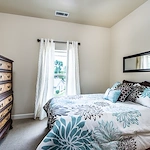 A cozy bedroom in Godfrey, Illinois, perfect for assisted living or memory care, with cream walls, a window with white curtains, a large bed with blue and brown floral bedding, a dark wood dresser on the left, and a mirror above the bed.