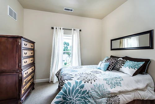 A cozy bedroom in Godfrey, Illinois, perfect for assisted living or memory care, with cream walls, a window with white curtains, a large bed with blue and brown floral bedding, a dark wood dresser on the left, and a mirror above the bed.