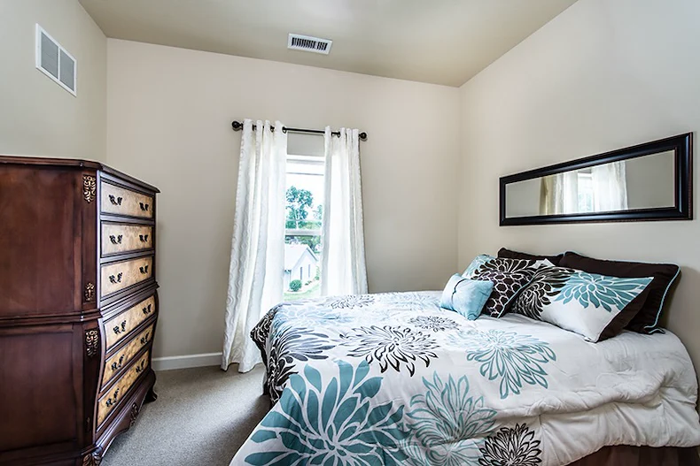 A cozy bedroom in Godfrey, Illinois, perfect for assisted living or memory care, with cream walls, a window with white curtains, a large bed with blue and brown floral bedding, a dark wood dresser on the left, and a mirror above the bed.