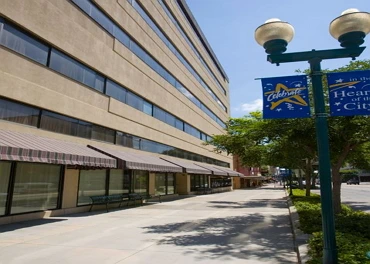 A sidewalk runs alongside a large beige building with striped awnings. A streetlamp with two globes and a blue “Celebrate” banner stands nearby. Trees line the sidewalk under a clear blue sky.