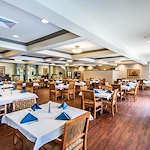 A spacious, well-lit dining room in Godfrey, Illinois, features wooden floors and tables with white tablecloths and blue napkins. Large windows, framed art, and potted plants create an elegant atmosphere ideal for assisted living or memory care residents.