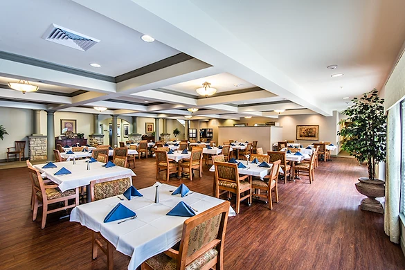 A spacious, well-lit dining room in Godfrey, Illinois, features wooden floors and tables with white tablecloths and blue napkins. Large windows, framed art, and potted plants create an elegant atmosphere ideal for assisted living or memory care residents.