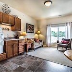 A cozy living area in Godfrey, Illinois offers assisted living comforts with a kitchenette featuring wooden cabinets, a sink, and microwave. Relax by the window or enjoy wall art that celebrates “Family,” “Friends,” and “Home.”.
