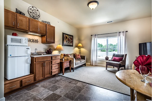 A cozy living area in Godfrey, Illinois offers assisted living comforts with a kitchenette featuring wooden cabinets, a sink, and microwave. Relax by the window or enjoy wall art that celebrates “Family,” “Friends,” and “Home.”.