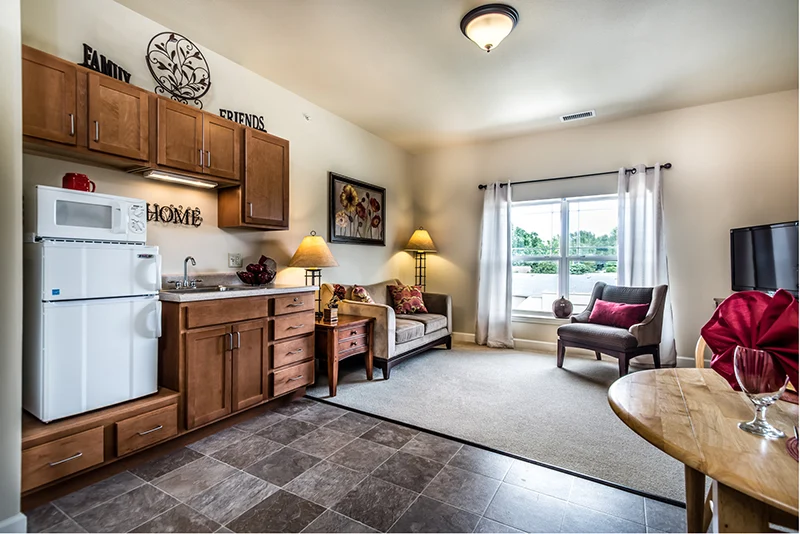 A cozy living area in Godfrey, Illinois offers assisted living comforts with a kitchenette featuring wooden cabinets, a sink, and microwave. Relax by the window or enjoy wall art that celebrates “Family,” “Friends,” and “Home.”.
