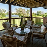 A covered wooden patio at Charter Senior Living features four wicker chairs with floral cushions around a glass-top table and potted plant. Overlooking a lawn, trees, and lake, it creates a peaceful outdoor setting for assisted living residents.
