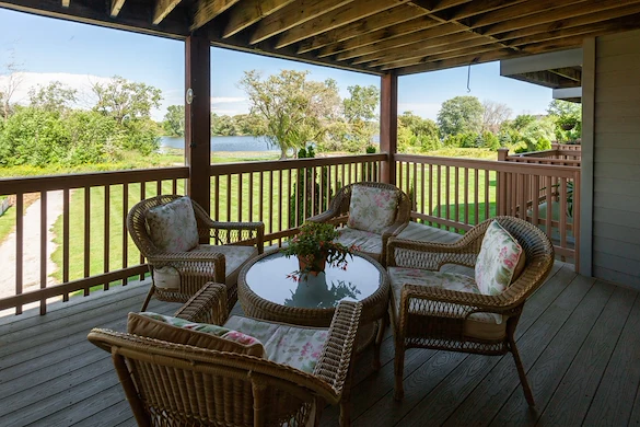 A covered wooden patio at Charter Senior Living features four wicker chairs with floral cushions around a glass-top table and potted plant. Overlooking a lawn, trees, and lake, it creates a peaceful outdoor setting for assisted living residents.