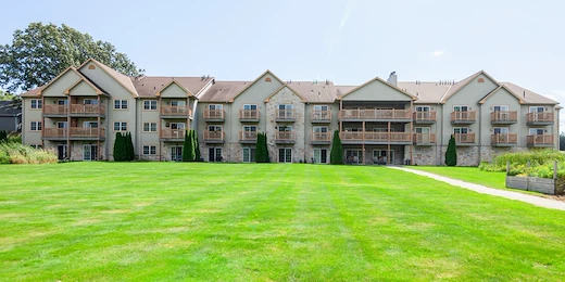 A wide, grassy lawn leads to a three-story assisted living apartment building with beige siding, stone accents, and balconies in Jackson, Wisconsin. Trees and shrubs frame the scene under a mostly clear sky with some sunlight.