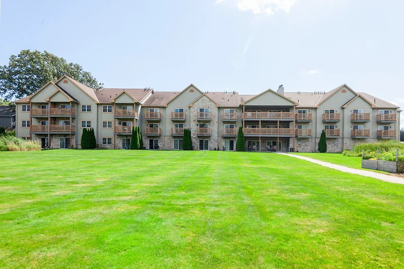 A wide, grassy lawn leads to a three-story assisted living apartment building with beige siding, stone accents, and balconies in Jackson, Wisconsin. Trees and shrubs frame the scene under a mostly clear sky with some sunlight.