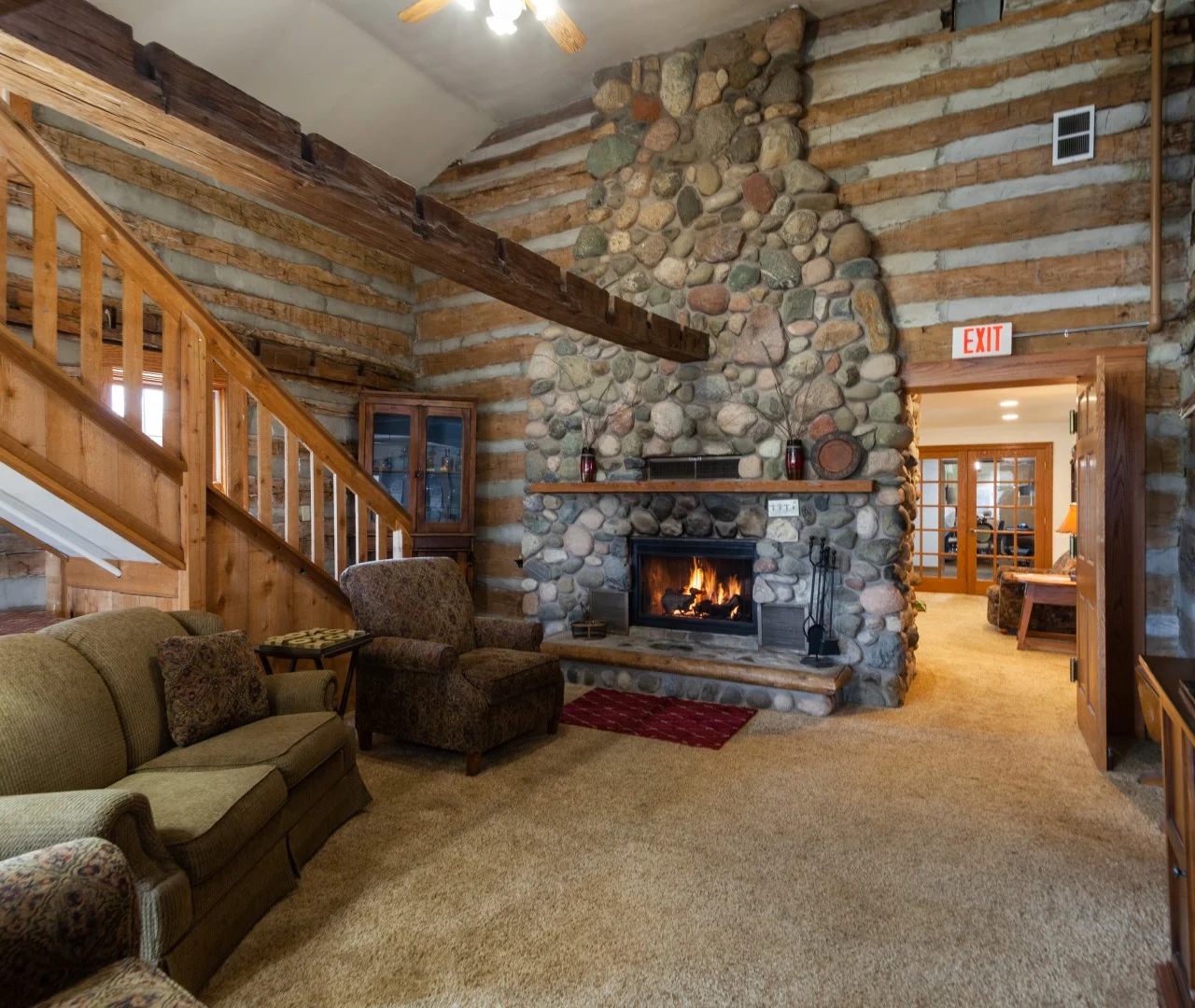 A cozy log cabin living room in Jackson, Wisconsin features a large stone fireplace with a burning fire, rustic wood beams, green sofa set, warm lighting, and two exit signs—an inviting space ideal for assisted living or memory care.