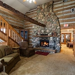 A cozy log cabin living room in Jackson, Wisconsin features a large stone fireplace with a burning fire, rustic wood beams, green sofa set, warm lighting, and two exit signs—an inviting space ideal for assisted living or memory care.