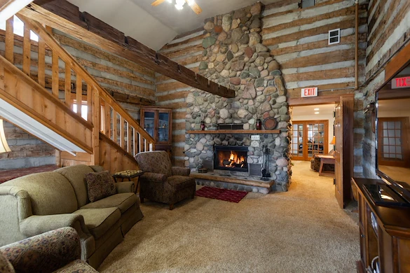 A cozy log cabin living room in Jackson, Wisconsin features a large stone fireplace with a burning fire, rustic wood beams, green sofa set, warm lighting, and two exit signs—an inviting space ideal for assisted living or memory care.