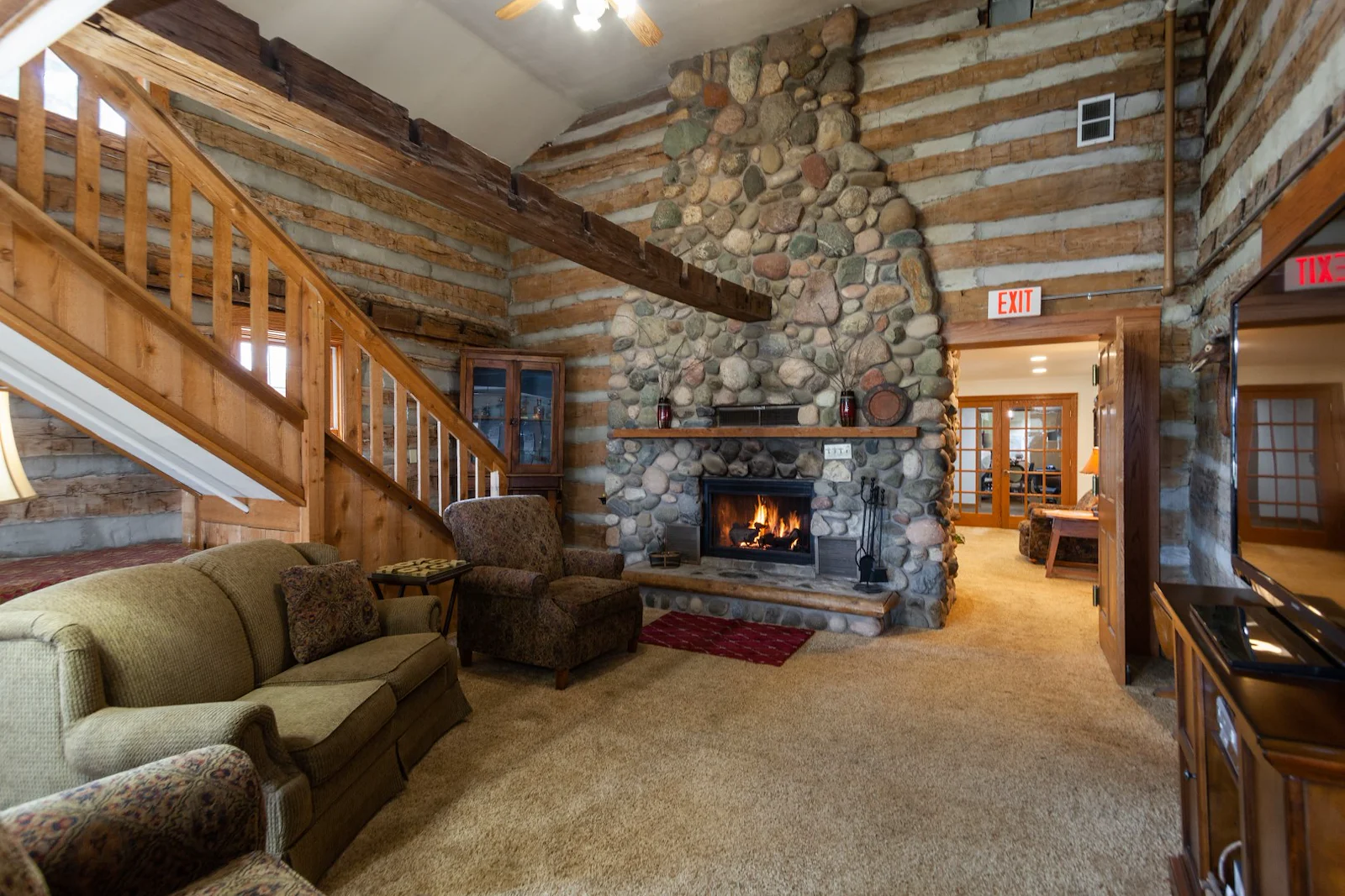 A cozy log cabin living room in Jackson, Wisconsin features a large stone fireplace with a burning fire, rustic wood beams, green sofa set, warm lighting, and two exit signs—an inviting space ideal for assisted living or memory care.