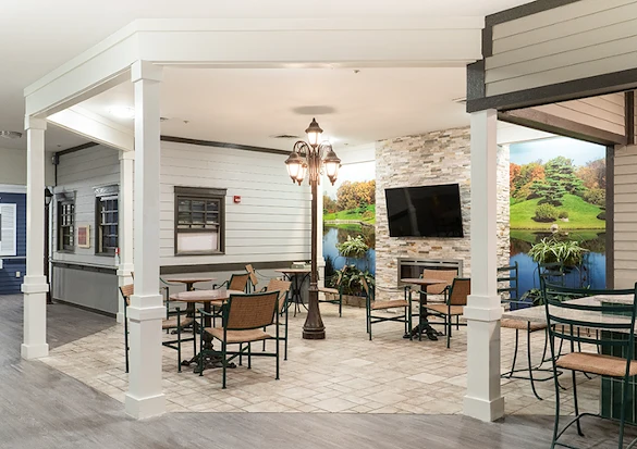 Cozy common space at Charter Senior Living of Hazel Crest in Hazel Crest, IL, with fireplace adorned by natural landscape wallpaper, TV, comfortable chairs, tables, and tile flooring, ideal for socializing and relaxing in assisted living and memory care.