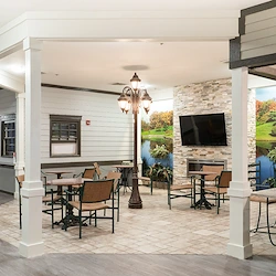 Cozy common space at Charter Senior Living of Hazel Crest in Hazel Crest, IL, with fireplace adorned by natural landscape wallpaper, TV, comfortable chairs, tables, and tile flooring, ideal for socializing and relaxing in assisted living and memory care.