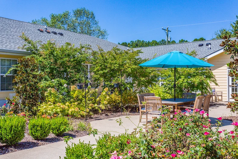Outdoor patio seating area with table, chairs, and umbrella at Charter Senior Living of Hermitage in Hermitage, TN, surrounded by vibrant flowers and greenery, offering a tranquil outdoor space for seniors in independent living, assisted living, and memory care.