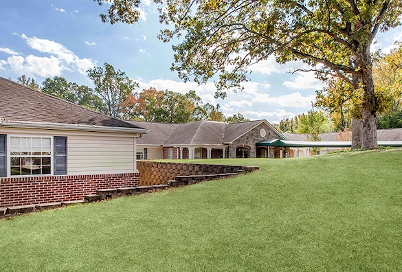 Another exterior view of Charter Senior Living of Jackson in Jackson, TN, featuring the brick building with covered awning and expansive grassy area with mature trees, offering assisted living and memory care in a peaceful setting.