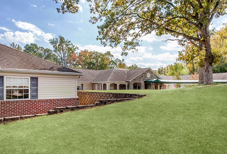 Another exterior view of Charter Senior Living of Jackson in Jackson, TN, featuring the brick building with covered awning and expansive grassy area with mature trees, offering assisted living and memory care in a peaceful setting.