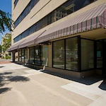 A city sidewalk runs alongside Crossroads House, a modern beige building with large windows and striped awnings in Lincoln, Nebraska. Shadows from trees fall across the pavement on a sunny day, reflecting the vibrant spirit of independent living.