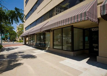 A city sidewalk runs alongside Crossroads House, a modern beige building with large windows and striped awnings in Lincoln, Nebraska. Shadows from trees fall across the pavement on a sunny day, reflecting the vibrant spirit of independent living.