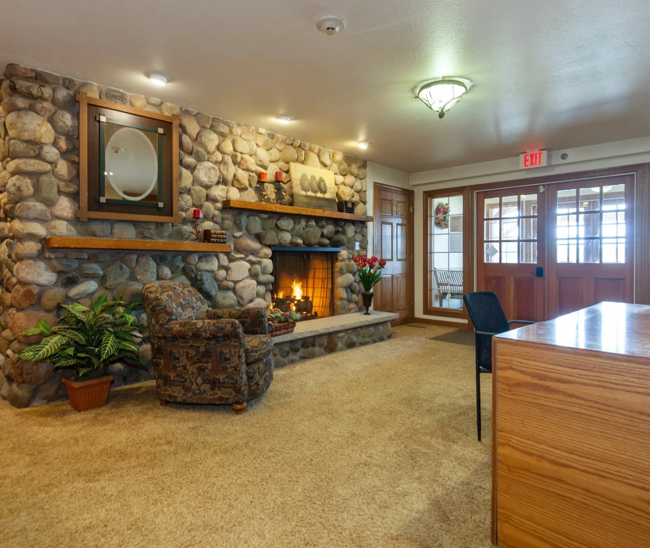 A cozy room at Charter Senior Living features a stone fireplace, lit fire, armchair, potted plant, and wooden desk. Glass doors and windows line the back wall. Warm lighting creates an inviting space ideal for assisted living or memory care residents.