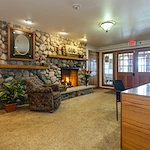 A cozy room at Charter Senior Living features a stone fireplace, lit fire, armchair, potted plant, and wooden desk. Glass doors and windows line the back wall. Warm lighting creates an inviting space ideal for assisted living or memory care residents.