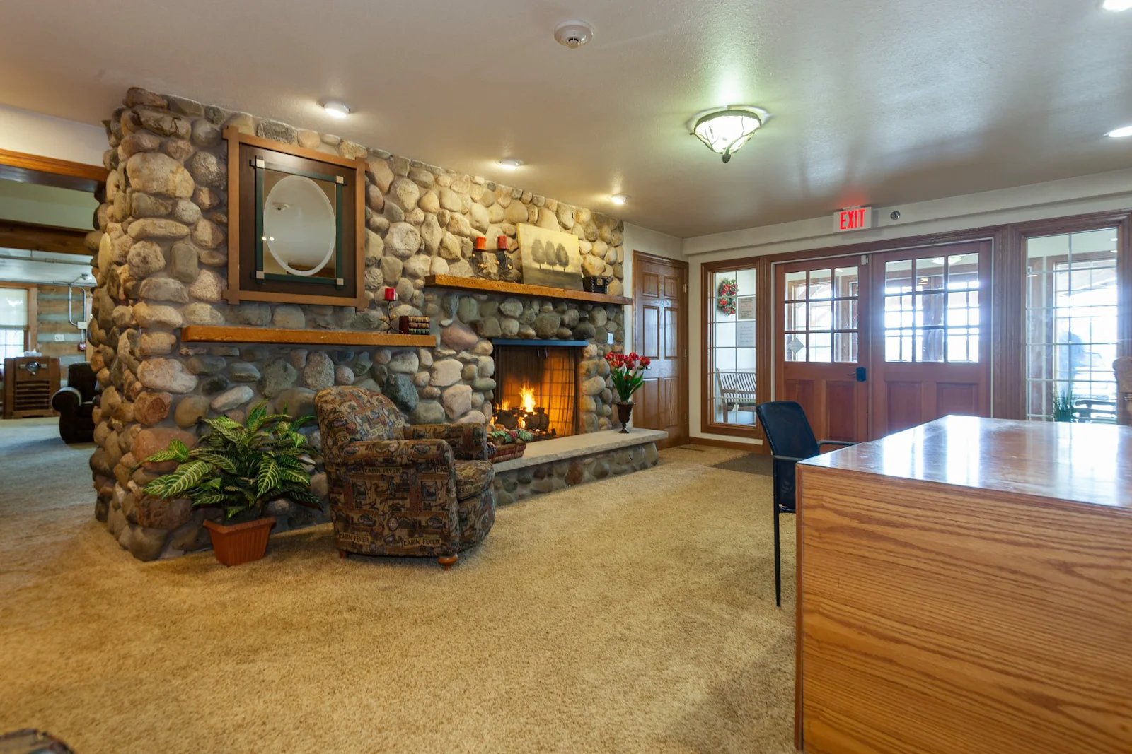 A cozy room at Charter Senior Living features a stone fireplace, lit fire, armchair, potted plant, and wooden desk. Glass doors and windows line the back wall. Warm lighting creates an inviting space ideal for assisted living or memory care residents.