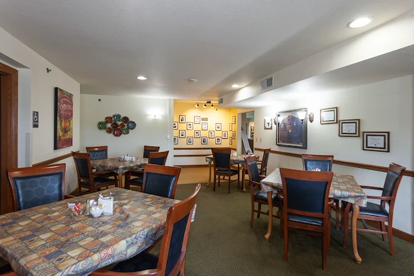 A dining room at Charter Senior Living in Jackson, Wisconsin features four wooden tables with patterned tablecloths and cushioned chairs. Framed photos and artwork adorn the walls, while overhead lights brighten the green-carpeted space.