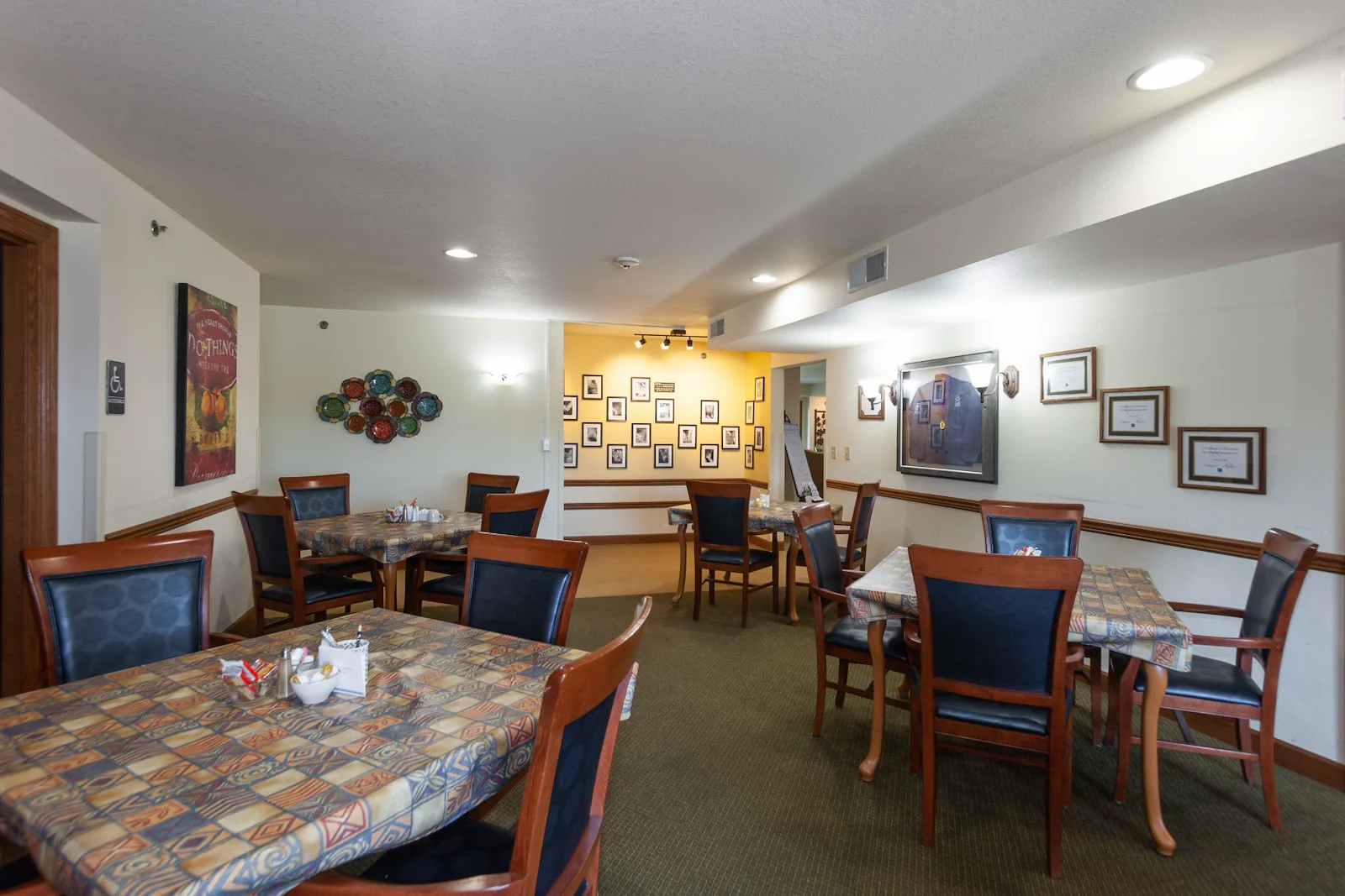 A dining room at Charter Senior Living in Jackson, Wisconsin features four wooden tables with patterned tablecloths and cushioned chairs. Framed photos and artwork adorn the walls, while overhead lights brighten the green-carpeted space.