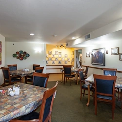 A dining room at Charter Senior Living in Jackson, Wisconsin features four wooden tables with patterned tablecloths and cushioned chairs. Framed photos and artwork adorn the walls, while overhead lights brighten the green-carpeted space.